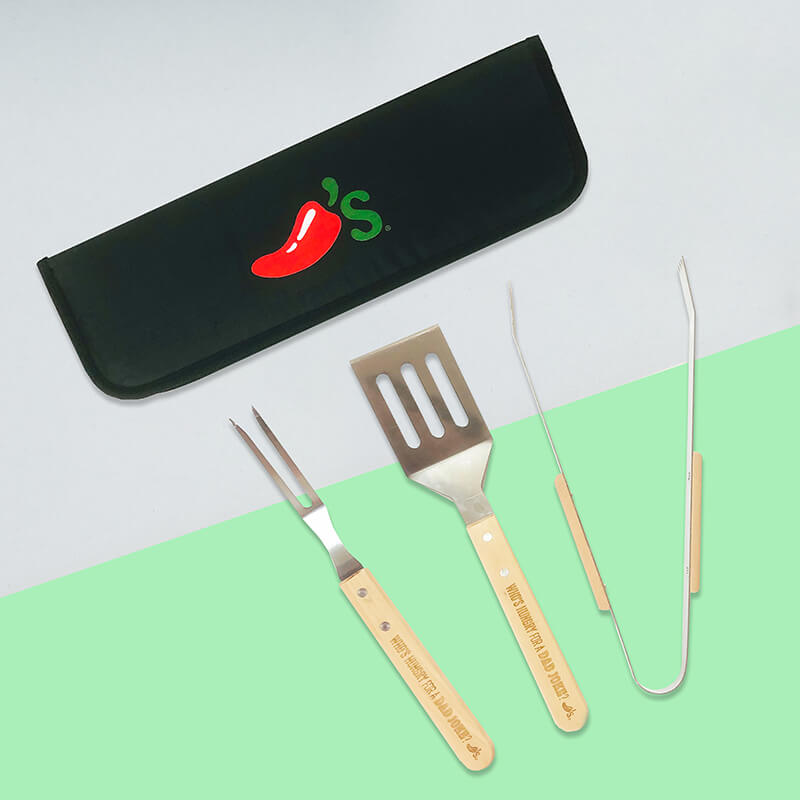 A flat lay of a grill set, with a black case with a bright red Chili's pepper with a green "'s" and a knife, fork, and tongs with "WHO'S HUNGRY FOR A DAD JOKE?" engraved on each handle.